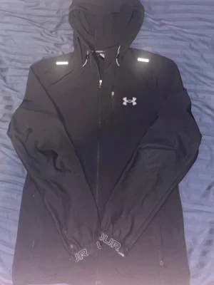 Svart vindjacka från Under Armour - Svart vindjacka med huva från Under Armour. Jackan har dragkedja framtill, bröstficka med dragkedja och reflekterande detaljer på axlarna. Justerbar huva med snören och elastiska muddar med logotypdetalj. Perfekt för blåsiga dagar.