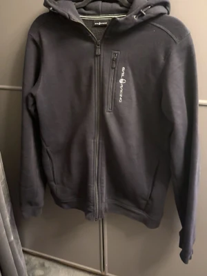 Svart hoodie från Sail Racing - Svart hoodie från Sail Racing med dragkedja framtill och en bröstficka med dragkedja. Märkets logga är tryckt i vitt på bröstet. Hoodien har justerbar huva med snören och ribbade muddar vid ärmslut och nederkant. Perfekt för en sportig och avslappnad stil.