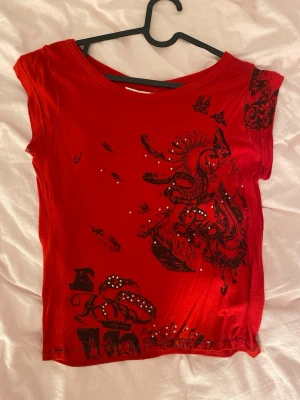Röd topp med svart tryck - Cool röd topp med svart grafiskt tryck och små glittriga stenar framtill. T-shirten har korta ärmar och rund halsringning. Materialet känns mjukt och stretchigt. Tröjan är omsydd av mig, de står storlek S i men eftersom jag sytt den mindre skulle jag mer säga XS.