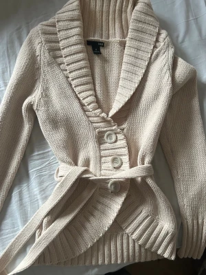  Kofta - Beige stickad kofta med bred sjalkrage, tre stora knappar framtill och matchande midjebälte. Långärmad modell med ribbade muddar och nederkant. Perfekt för lager på lager och en avslappnad men snygg look.