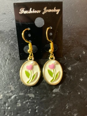 Guldiga örhängen med blommotiv - Söta guldiga örhängen med oval form och hängande design. Framsidan har ett motiv av en rosa blomma med gröna blad på vit bakgrund. Perfekta för att ge en färgklick till din stil.