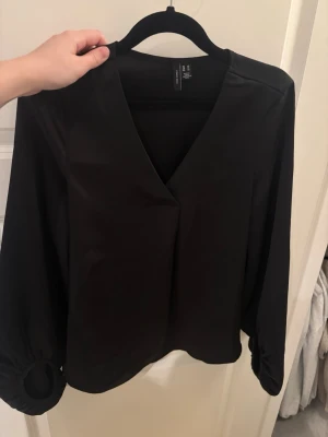 Svart blus med v-ringning från VERO MODA - Stilren svart blus från VERO MODA med v-ringning och långa puffärmar som har dekorativa rynkade detaljer vid ärmsluten. Tillverkad i ett glansigt, mjukt tyg som ger en elegant känsla. Perfekt att matcha med både jeans och kjol.