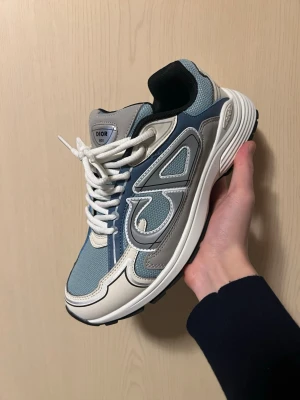 Dior B30 ljusblåa - Säljer ett par Dior B30 sneakers i blått, beige och vitt med coola detaljer och reflekterande inslag. Ovandelen är i mesh och läder med tydlig Dior-logga på sidan. Skorna har vita snören och en chunky sula som ger en modern vibe. Skick: 9/10, aldrig använda. 