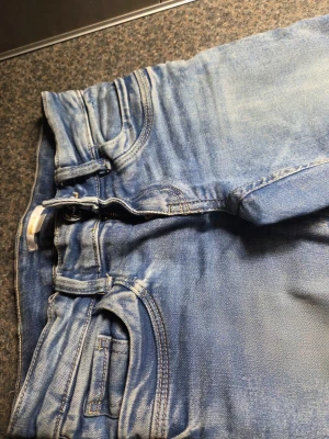 Blå bootcut jeans - Säljer ett par klassiska blå jeans med raka ben och lätt slitna detaljer. Jeansen har fem fickor, knapp- och dragkedjeförslutning samt ljus tvätt. Perfekt för dig som gillar en avslappnad och tidlös look.