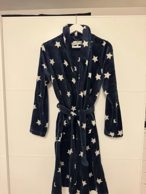 Mjuk marinblå morgonrock med stjärnor - Supermjuk marinblå morgonrock med vita stjärnor, två stora fickor framtill och knytskärp i midjan. Perfekt för att chilla hemma och hålla sig varm. Tillverkad i fluffigt fleece-material som känns riktigt skönt mot huden.😇