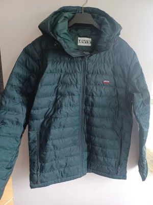 Mörkgrön jacka från Levi's - Snygg mörkgrön pufferjacka från Levi's med huva och dragkedja framtill.  Jackan har två sidofickor med dragkedja och diskret Levi's-logga på bröstet. Perfekt för kalla dagar och tillverkad i lätt, quiltat material som håller värmen. Använd bara några ggr. Mycket bra skick.