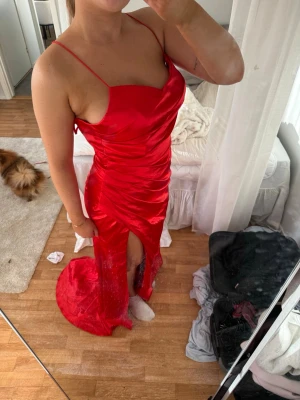 Röd långklänning med snörning - Säljer en röd långklänning med smala axelband och snygg snörning i ryggen. Klänningen har en slits framtill och glittriga detaljer längst ner. Sitter figurnära och har en elegant, svepande form. Perfekt för bal eller festliga tillfällen.
