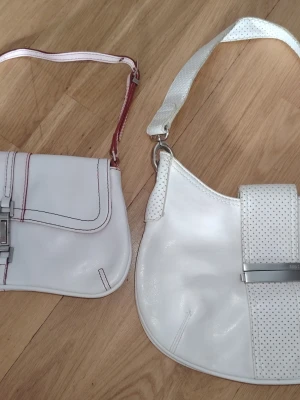 Vit handväska  från Mango med metallspänne - Två vita handväskor från Mango. Den ena väskan har röda sömmar och en röd rem sat röda detaljer, den andra har perforerade detaljer på remmen och framsidan. Båda väskorna har metallspännen med MNG-logotypen. Den större väskan har ett grått innerfoder med flera fack, medan den mindre väskan har ett rött innerfoder med ett fack.  Den vita är något mer använd och ha fler defekter/skavanker (se bilder). Den lilla vit/röda väskan har knappt någon defekt, syns bara en mindre dov märke på framsidan som