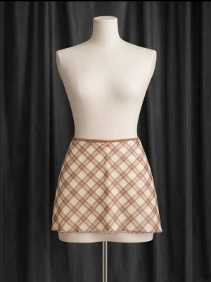 Beige rutig minikjol med volangkant - Supersöt beige minikjol med rutigt mönster i bruna toner och liten volangkant längst ner. Kjolen har en enkel, rak passform och är perfekt för dig som gillar en trendig och ungdomlig stil.