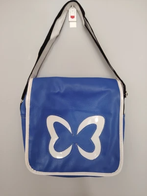 Blå axelväska med fjärilsmotiv - Cool blå axelväska i konstläder med vit kant och stort vitt fjärilsmotiv på framsidan. Väskan har justerbar axelrem i svart och vit. Har ett stort fack på insidan. Perfekt för dig som vill sticka ut med en färgglad och unik accessoar. Funkar utmärkt till plugget eller till vardags. 
