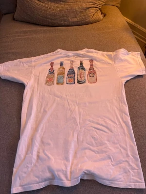 2 St BVALDI T-shirt (Nyskick) - Säljer två t-shirts: en vit med färgglada flaskor på ryggen och en svart med ett kreditkorts-inspirerat tryck i glittriga stenar på bröstet. Tröjorna är använda endast någon gång.