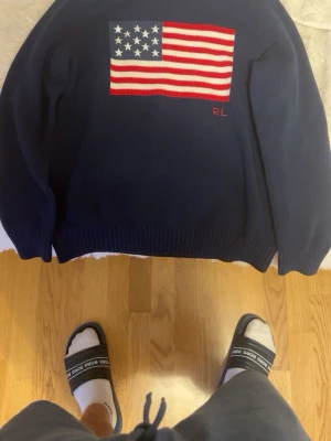 Mörkblå stickad tröja Ralph Lauren - Snygg mörkblå stickad tröja från Ralph Lauren med amerikansk flagga broderad på bröstet och RL-detalj i rött. Klassisk rund halsringning och ribbade muddar vid ärmslut och nederkant. Perfekt för dig som gillar preppy stil.