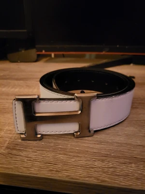 Vitt H bälte  - Snyggt vitt skärp med ett stort metallfärgat H-spänne. Bältet har svarta kanter och är gjort i skinn.