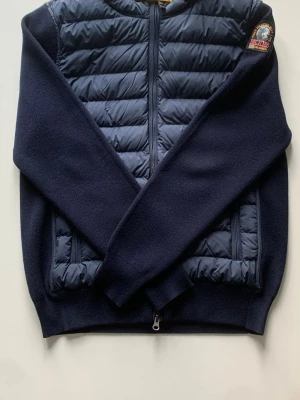 Parajumpers Illuga Cardigan - Säljer en snygg mörkblå cardigan från Parajumpers i storlek XS. Jackan har quiltad framsida och huva, stickade ärmar och rygg samt dragkedja framtill. Klassisk patch på ärmen och gul detalj vid halsen. Perfekt nu inför våren. Cardiganen är i nyskick, bara använd ett fåtal gånger. Skickas samma dag. Allt medföljer såsom box, brev och tags. Pris kan diskuteras. Kontakta vid intresse.