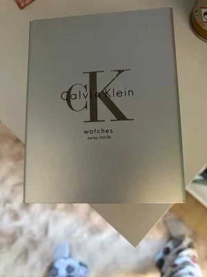 Calvin Klein silverfärgad klocka - Snygg och stilren klocka från Calvin Klein med rektangulär svart urtavla och silverfärgat armband i metall. Klockan har minimalistisk design och diskret CK-logga på urtavlan. Perfekt accessoar för dig som gillar klassisk och modern stil.