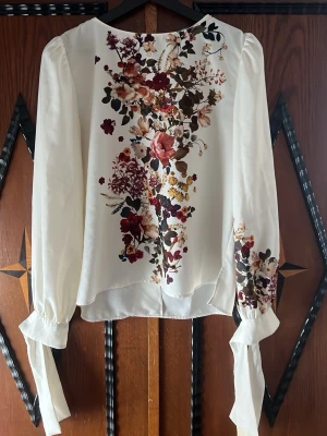 Vit blus med blommigt tryck och knytband - Stilren vit blus med färgstarkt blommönster framtill och på ärmarna. Långa ärmar med puff och breda knytband vid handlederna ger en trendig look. Tillverkad i ett lätt, glansigt material som faller snyggt. Perfekt för dig som vill sticka ut med detaljer.