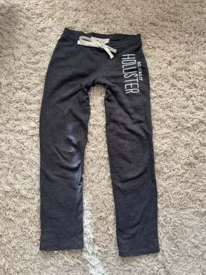 Mörkgrå mjukisbyxor från Hollister - Mörkgrå Lågmidjade mjukisbyxor från Hollister med vit dragsko i midjan och tryckt logga på ena benet. Sköna och raka ben, perfekt för chill eller träning. Mjukt material som känns bekvämt hela dagen.
