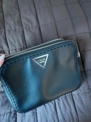 Svart handväska från Guess - Nästan helt oanvänd svart handväska från Guess i slätt konstläder med silverfärgad dragkedja och logga framtill. Väskan har ett ljust innerfoder med Guess-mönster och en innerficka med dragkedja. Tyvärr ingår ingen axelband, annars som ny🌸