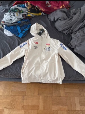 Vit vindjacka från Umbro Santos FC - Snygg vit vindjacka från Umbro med Santos FC-logga på bröstet och flera tryckta sponsorloggor i rött och blått. Jackan har huva, dragkedja framtill och är tillverkad i lätt material som passar perfekt för blåsiga dagar.