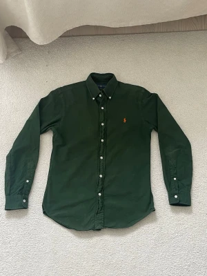 Mörkgrön skjorta från Ralph Lauren - Klassisk mörkgrön skjorta från Ralph Lauren med orange broderad logga på bröstet. Skjortan har långa ärmar, knappar hela vägen och button-down krage. Tillverkad i mjuk bomull för en bekväm känsla och snygg passform.