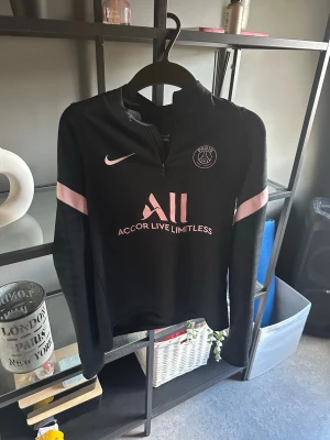 Svart PSG träningströja Nike - Svart långärmad träningströja från Nike och PSG med rosa detaljer och halv dragkedja. PSG-logga och Nike Swoosh på bröstet, samt tryck med 'Accor Live Limitless' och 'Visit Rwanda'. Perfekt för fotbollsträning eller chill.