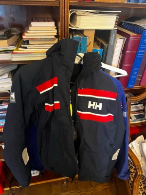 Helly Hansen marinblå vindjacka - Snygg marinblå vindjacka från Helly Hansen med röda och vita ränder på bröstet och ärmen. Jackan har hög krage, huva och dragkedja framtill. Perfekt för blåsiga dagar och har tydlig HH-logga både fram och bak. Storlek Xl men känns mer som (L-M)