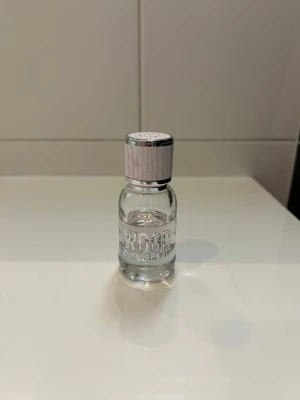 Dsquared2 Wood parfym mini - En liten flaska Dsquared2 Wood parfym med transparent glasflaska och ljusrosa kork med silverdetaljer. Korken har ett lönnlövsmotiv på toppen. Perfekt att ha med i väskan eller på resan.