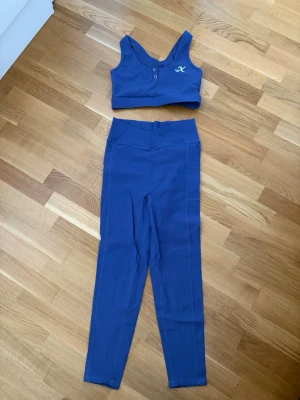 Blått träningsset med sport-bh och tights - Snyggt blå-lila träningsset från Xshapes med matchande sport-bh och höga tights. Sport-bh:n har knappar framtill och logga på bröstet, tightsen har hög midja, fickor på sidorna, scrunch-detalj bak och logga framtill. Perfekt för gym eller yoga.