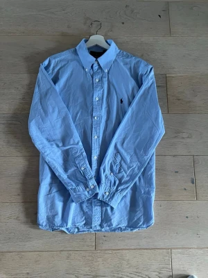 Blå skjorta från Ralph Lauren - Klassisk ljusblå skjorta från Ralph Lauren med button-down krage och broderad logga på bröstet. Skjortan har lång ärm, vita knappar och är tillverkad i mjuk bomull. Perfekt för en clean och stilren look.