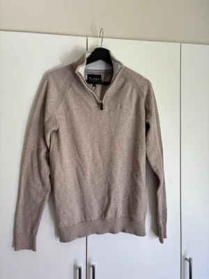 Beige stickad tröja med halv dragkedja - Stickad beige tröja från Bondelid med halv dragkedja och hög krage. Tröjan har ribbade muddar vid ärmslut och nederkant samt en stilren, enkel design. Perfekt för lager-på-lager och passar till många olika outfits.