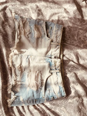 Ljusblå slitna jeansshorts med fransar - Säljer ett par ljusblå jeansshorts med slitna detaljer och fransig kant. Shortsen har hög midja, klassiska fickor fram och bak samt en cool, avslappnad vibe. Perfekta för varma dagar och festivaler.