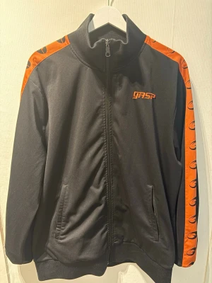 Svart träningsjacka från GASP - Svart träningsjacka med dragkedja från GASP. Jackan har orangea ränder med logotyp längs axlar och ärmar samt broderad GASP-logga på bröstet. Hög krage och två fickor framtill. Perfekt för gymmet eller sportaktiviteter.