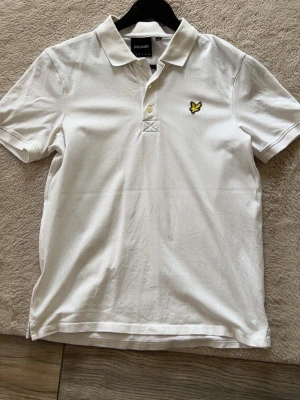 Vit pikétröja från Lyle & Scott - Klassisk vit pikétröja från Lyle & Scott med gul logga på bröstet. Tröjan har krage, knappar framtill och är kortärmad. Perfekt för en clean och stilren look. Mycket bra skick.
