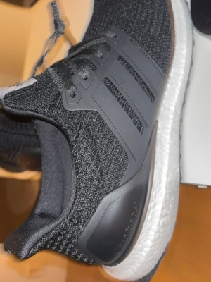 Adidas Ultraboost svarta sneakers - Svarta Adidas Ultraboost sneakers med stickad ovandel och klassiska tre ränder på sidan. Skorna har en vit Boost-sula för extra komfort och svart hälkappa. Perfekta för dig som gillar sportig och stilren look.