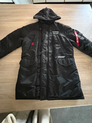 Svart parkas från Alpha Industries - Säljer en svart parkas från Alpha Industries med huva och dragkedja samt knappar framtill. Jackan har flera fickor, tydlig 'Remove Before Flight'-tag på ärmen och vita tryckdetaljer. Tillverkad i slitstarkt syntetmaterial, perfekt för kalla dagar.