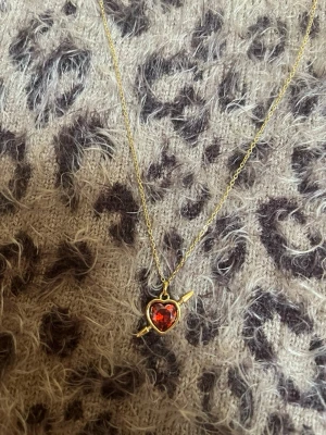 Guldhalsband med röd hjärtberlock spjut - Sött halsband i guldton med tunn kedja och en hjärtformad berlock med röd sten. Berlocken har en pil genom hjärtat vilket ger en unik och lekfull look. Perfekt accessoar för att lyfta din outfit.rostfri 