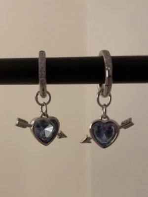 Hjärthängande örhängen med blå hjärtan silver - Snygga örhängen i silverfärg med hjärtformade hängen och blå glittrande sten. Hängena har en pil genom hjärtat för en unik touch. Perfekta för dig som gillar detaljer och vill sticka ut lite extra. Rostfri 