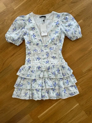  Slutsåld student Blommig kortklänning från Zara - Superfin kortklänning från Zara med blommigt mönster i blått och lila på vit botten. Klänningen har volanger, v-ringning och puffärmar. Tillverkad i ett lätt bomullstyg, perfekt för varma dagar. Söt spetsdetalj längs urringningen och midjan.