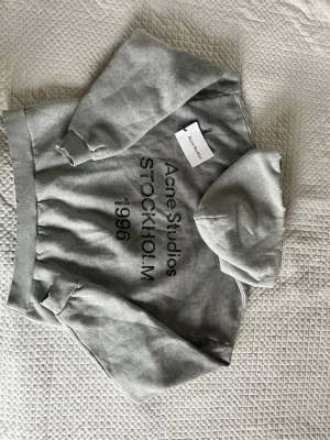 Grå hoodie från Acne Studios - Snygg grå hoodie från Acne Studios med svart tryck på framsidan där det står 'Acne Studios STOCKHOLM 1986'. Klassisk passform med huva och ribbade muddar vid ärmslut och midja. Perfekt för en chill och stilren look. Den är äkta köpt för 4700kr
