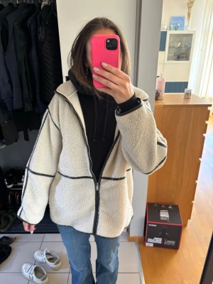 Vit teddyjacka med svarta detaljer - Säljer en oversized vit/beige teddyjacka med svarta/brun kontrastsömmar och dragkedja framtill. Jackan har en mysig, fluffig känsla och är perfekt för lager-på-lager. Passar dig som gillar streetstyle och vill ha en chill look. Prislappen är på och varan är aldrig använd!