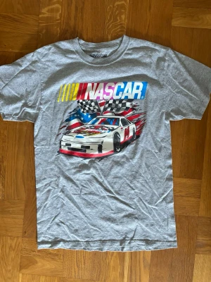 Grå NASCAR t-shirt vintage  - Snygg grå t-shirt från NASCAR med stort färgglatt tryck av en racingbil, amerikansk flagga och rutiga flaggor på bröstet. Klassisk passform och rund halsringning. Perfekt för dig som gillar motorsport och streetstyle. Size S