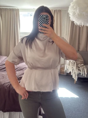 Ljusgrå satinblus med peplum - Snygg begie blus i satin med korta ärmar och peplum-modell. Blusen har hög hals och en lätt glansig finish som ger en trendig look. Perfekt att styla med jeans eller kjol för en chic outfit.