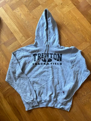 Grå hoodie Trenton Track & Field - Snygg grå hoodie från Champion med trycket 'TRENTON TRACK & FIELD' på framsidan och 'MOORE' på ryggen. Klassisk modell med huva och dragsko samt ribbade muddar. Perfekt för en sportig och avslappnad stil. Storlek M, fint skick! 