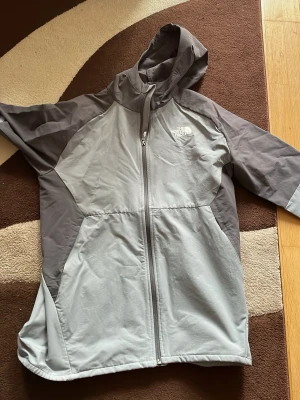 Grå vindjacka från The North Face - Snygg grå vindjacka från The North Face med huva och hel dragkedja framtill. Jackan har två olika grå nyanser och logga på bröstet. Perfekt för blåsiga dagar och outdoor-aktiviteter. Tillverkad i lätt och slitstarkt material.