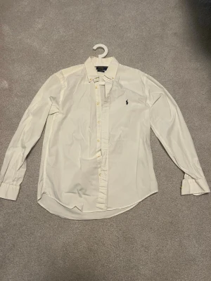 Vit skjorta från Ralph Lauren - Klassisk vit skjorta från Ralph Lauren med broderad logga på bröstet. Skjortan har lång ärm, knappar framtill och krage. Perfekt för en clean och stilren look.