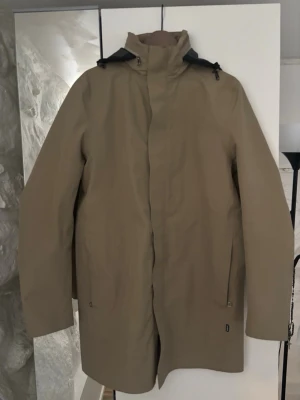 Peak performance regnjacka med huva - Snygg beige regnjacka med hög krage och justerbar huva. Jackan har dragkedja och tryckknappar framtill samt två sidofickor. Tillverkad i vattenavvisande material, perfekt för regniga dagar. Enkel och stilren design som passar till det mesta.
