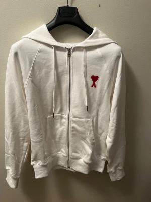 Vit zip-hoodie från Ami Paris - Snygg vit hoodie med dragkedja från Ami Paris. Klassisk design med huva, dragsnören och känguruficka. På bröstet finns Ami:s ikoniska röda hjärta med A. Perfekt för en clean och stilren look.