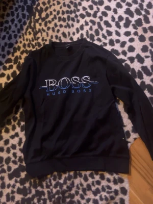 Svart sweatshirt från Hugo Boss - Svart sweatshirt med rund hals och stort BOSS-tryck i vitt och blått framtill. Slim fit-modell med ribbade muddar vid ärmslut och nederkant. Perfekt för en stilren och avslappnad look.