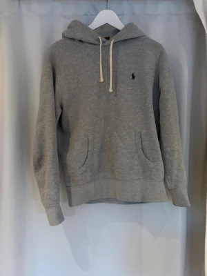 Grå hoodie från Polo Ralph Lauren - Klassisk grå hoodie från Polo Ralph Lauren med broderad logga på bröstet. Hoodien har huva med vita snören och en stor magficka framtill. Tillverkad i mjukt bomullsmaterial och har ribbade muddar vid ärmslut och nederkant.