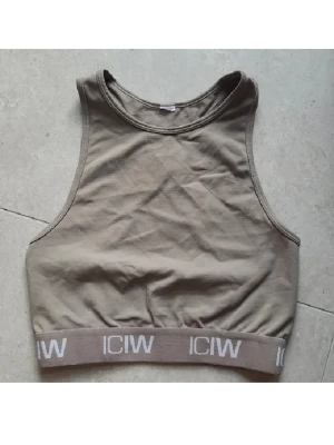 Beige topp från ICIW - Snygg beige sporttopp eller sportbh från ICANIWILL med bred resår nertill där loggan syns i vitt. Modellen är ärmlös med hög halsringning och brottarrygg. Perfekt för träning eller chill dagar. Mjuk bomullskänsla och stilren design.